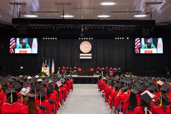 View down center aisle on the ceremony floor shows graduates seated in rows on both sides and Dean Delva on two jumbotron screens