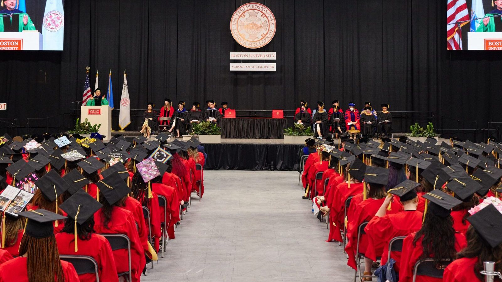 View down center aisle on the ceremony floor shows graduates seated in rows on both sides and Dean Delva on two jumbotron screens
