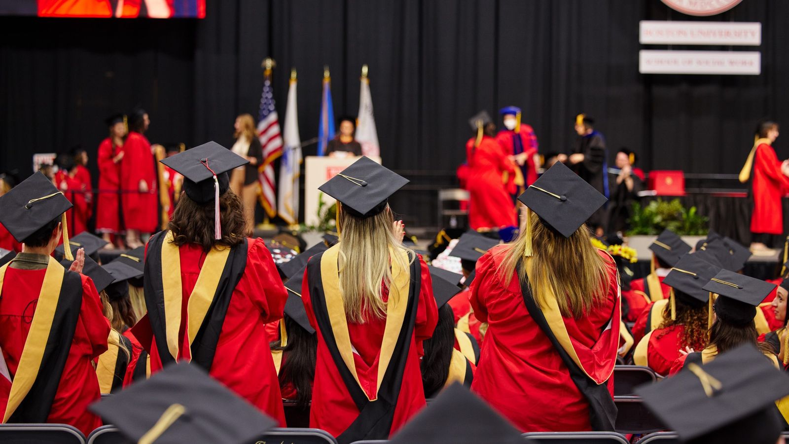 Hooded graduates stand at seats as graduate on stage receives diploma