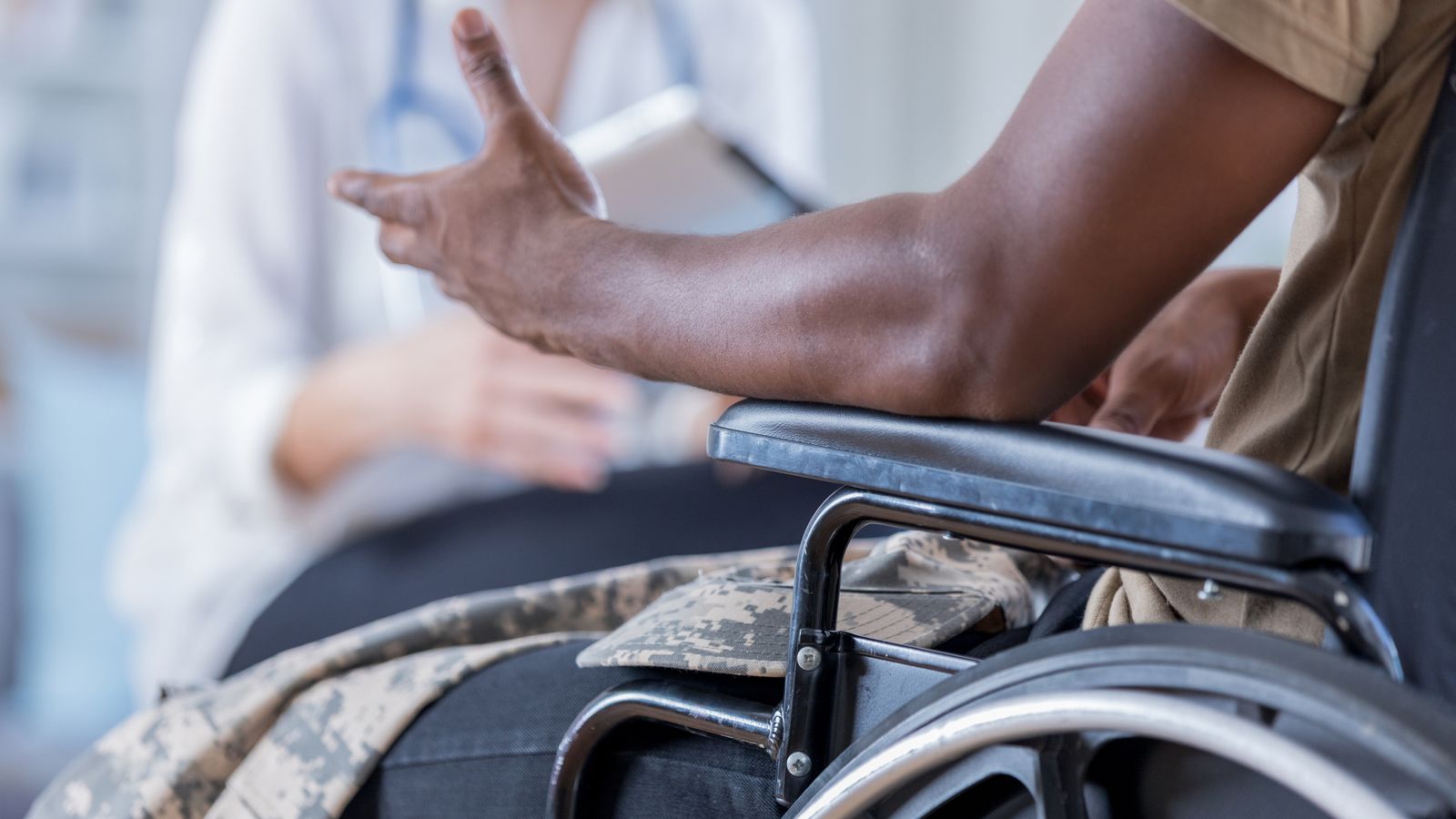 In this closeup, both a doctor and military patient are unrecognizable as the patient shares his symptoms. Focus is on the wheelchair bound officer as he gestures during the discussion.