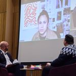 Photo of the event moderator and author on stage looking at photographer who is speaking on screen via Zoom