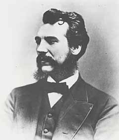 Black and white photo of Alexander Graham Bell as a young man. Bell is pictured with longish hair on head, and connecting beard and moustache. He wears a suit with vest over a white shirt, a black bowtie. The background of the photo is faded grey around him and fades to white toward the edges of the image.