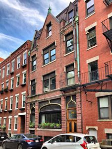 Color image modern day of the building at 36 Hull Street where the medical mission was located in the 19th century. The building has a brick facade with a peaked top and small windows on three floors. The bottom floor features a large picture window with flower box across it. Cars are parked on the street in front of the building. The brick building is adjacent to the other brick buildings.