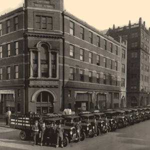 Black and white image of a large brick building with a line of trucks with men and women in front of the building. Building is approximately six stories. Doorways are arched. Windows are small and in long rows of 8.