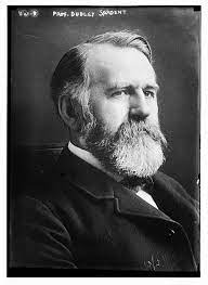 Black and white Image of Dudley Sargent. Image of man with beard and mustache and short men's haircut wearing a black suit with white shirt. Bears is mostly white but some grey and black hairs are dispersed through moustache and head. Grey background and sargent's name appears above his image