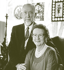 Image of Dr. Albert Danielsen and his wife, Jessie, Image is black and white with Albert standing on the left side of the photo, and wife sitting on the right. Albert is wearing a solid colored suit with stripped tie, and Ms. He has a thin face with receding hairline, and white goateeDanielsen is wearing a round neck top. She wears glasses, smiling, and has tight curly hair.