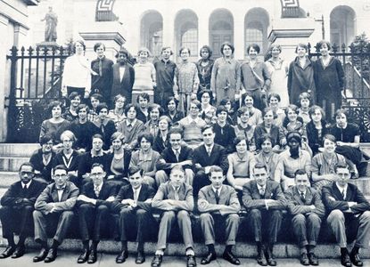 Class of 1918, black and white photo of approximately 60 students sitting in rows on steps of a building with three arches behind them. All in fashion from 1918 period. there is a mix of women and men.