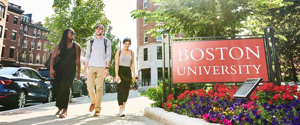 Students walking along a sidewalk next to a large Boston University sign