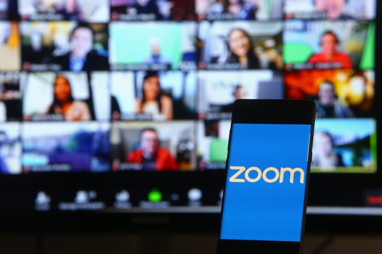 A phone showing the Zoom logo in front of a laptop screen showing many people in a video conference