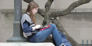 A young woman sits on a step in front of a tree and light pole as she works on her laptop.