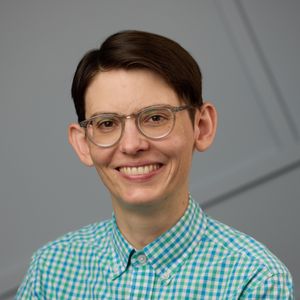 Allison Reilly's headshot. Photo of a smiling person from their shoulders up. They have short brown hair, clear-rimmed glasses, peach/white skin and are wearing a green and blue checkered shirt. The background behind them is gray with intersecting lines.