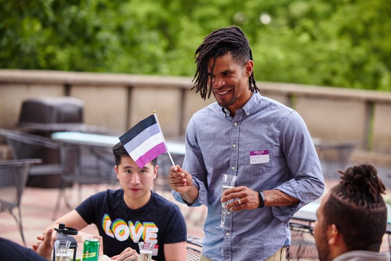 A man in a button up holding a small asexual flag (black, grey, white, and purple stripes) in one hand and a glass of Prosecco in the other. Another man is sitting behind him watching.