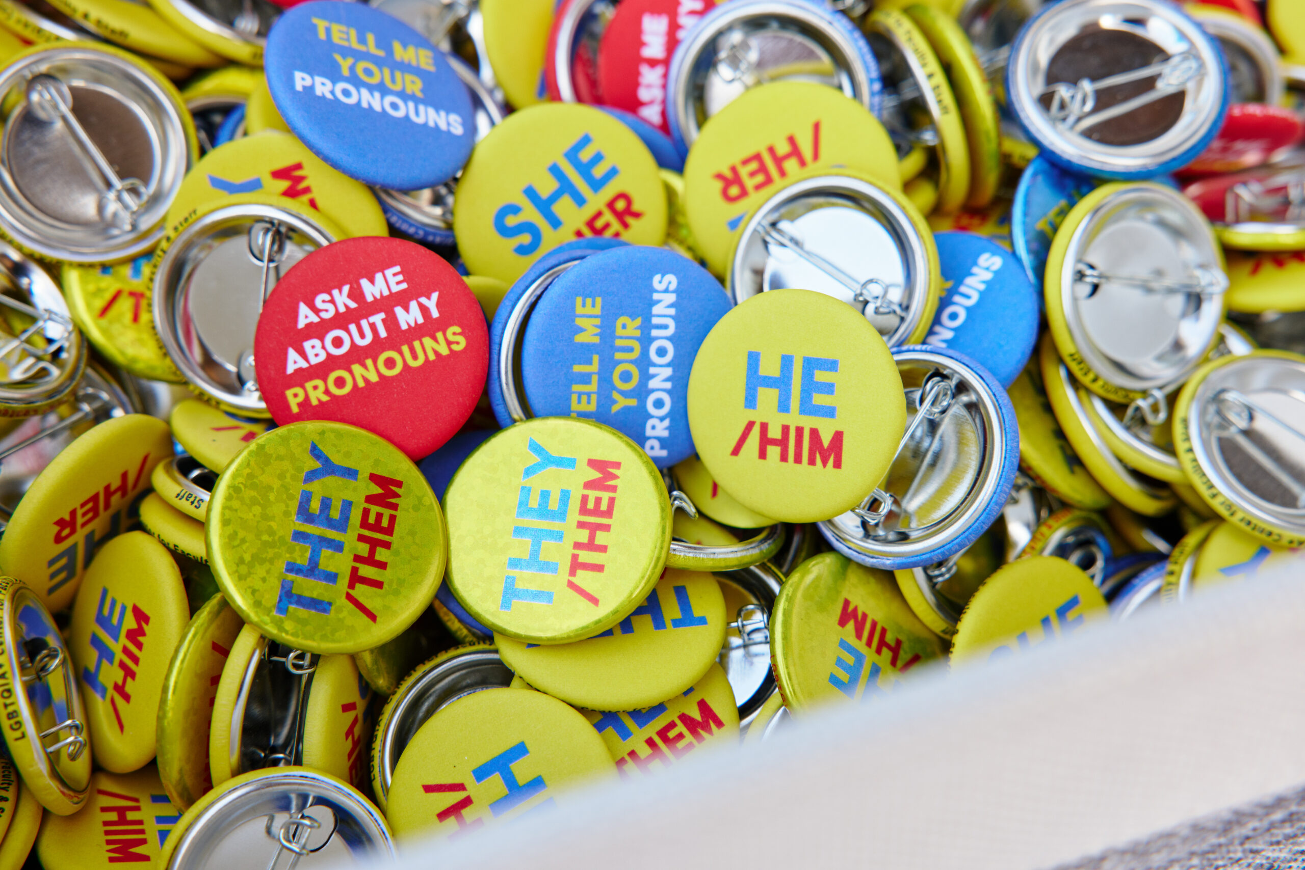 A close up photo of a variety of pronoun pins with the following pronouns: he/him, they/them, Ask me about my pronouns, She/Her, Tell me about your pronouns. The pins are jumbled up in a basket.