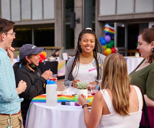 A group of people standing around a cocktail table laughing and enjoying conversation. The table has a rainbow table runner with a water bottle, plates of food, and cocktails on the table.