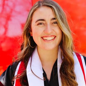 A headshot of Shelby Cattani. She has light skin, with long wavy blonde hair and is wearing a black gown wtih white and red stole. Shelby is standing in front of a red background.