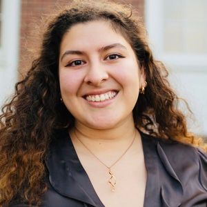 This is an image of Kayla. She has light skin, curly brown hair, and brown eyes. She is wearing a grey blouse and has gold earrings and a gold necklace.