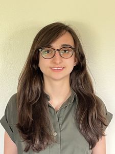 This is an image of Beatrice. She has long brown hair and fair skin. She is wearing glasses and a forest green/ grey shirt.