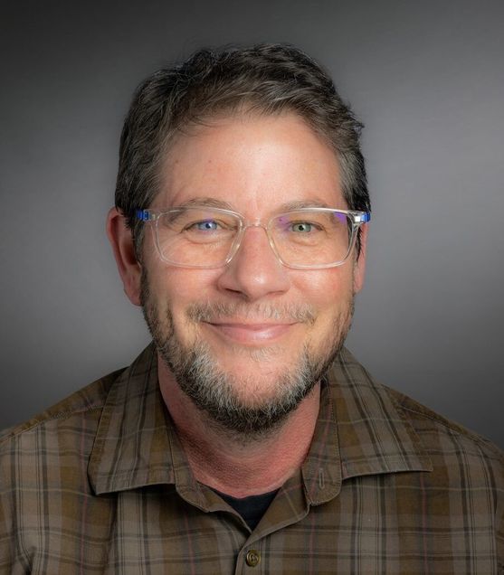 A photo of Chris Hinesley, Associate Director of the LGBTQIA+ Center for Faculty & Staff. The photo is of a man with light peach-colored skin, short hair, medium beard, wearing a brown plaid shirt and glasses.
