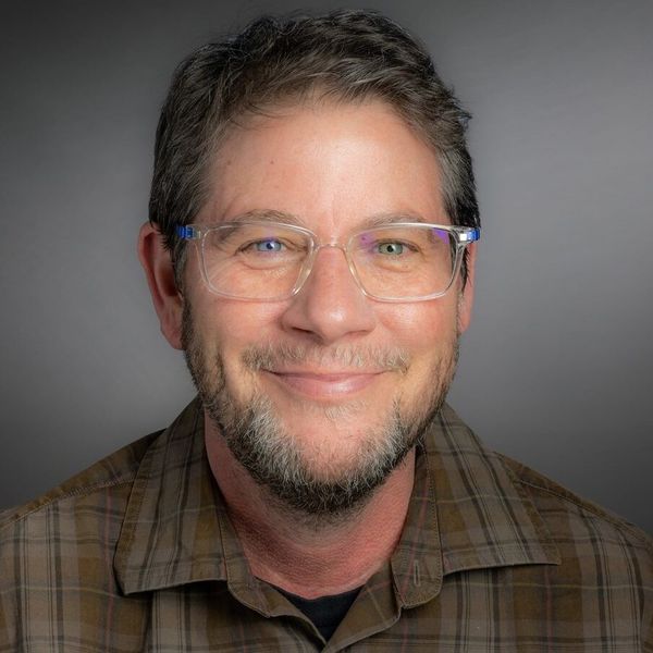 Chris Hinesley, Associate Director of the LGBTQIA+ Center for Faculty & Staff. with short hair, wearing a brown plaid shirt and glasses.
