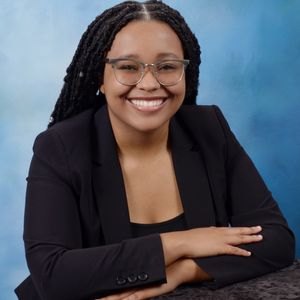 Headshot of Caryne A. Nicholas. They have brown skin and dark brown hair in long locs, parted in the middle. They are wearing a black blazer, and brown frame glasses. They have a huge smile on their face, while crossing their arm on a surface. The background is blue.