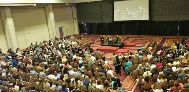 Overhead view of a large room with seated audience. Dr. Beverly Tatum sits on a stage with Dr. Michelle Porche.