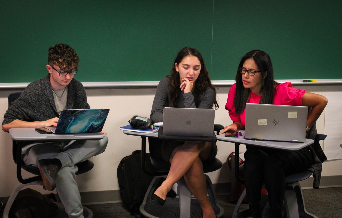 Three students huddled together in class talking in front of a green chalk board