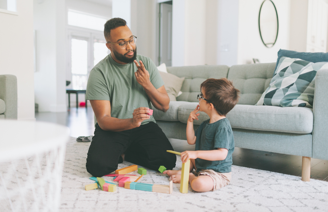 man sits on carpet in front of light blue couch using ASL to communicate to small child sitting next to him