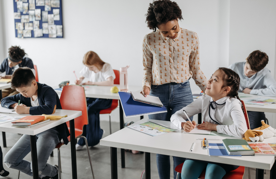 Shows a teacher standing over a student at a desk with other students at desks