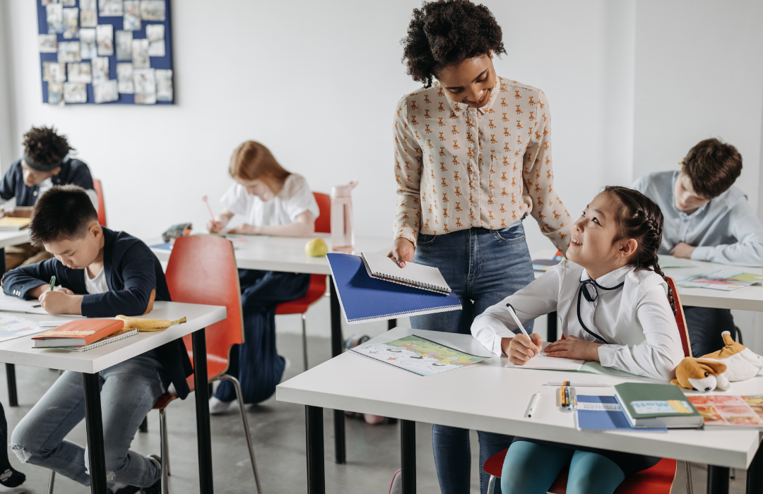 Shows a teacher standing over a student at a desk with other students at desks