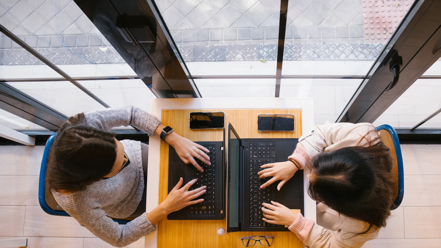 A picture of two young women sitting across from each other at a desk. Both of them are typing on laptops.