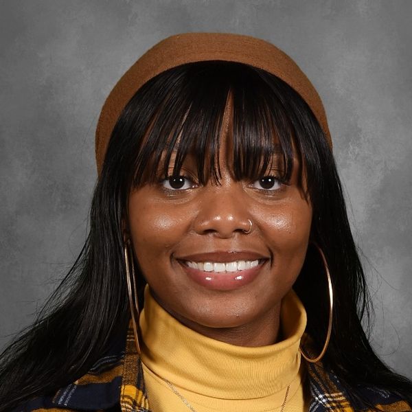 Shows Jacqueline Ferguson, who has long black hair and is wearing a yellow turtleneck and plaid shirt