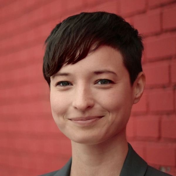 Shows Maya Bialik, head and shoulders against a red background, short brown hair and a green jacket