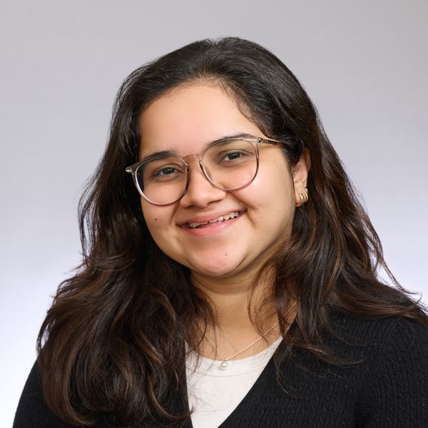 Shows Mitali Termurnikar, who has shoulder length dark hair, is wearing glasses, a white top, and a black v neck sweater