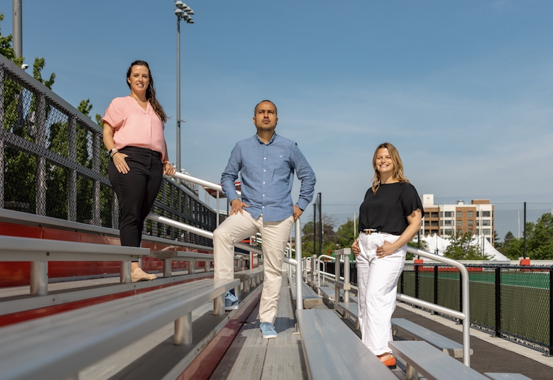 From L-R Anna Ward, Edson Filho, Carly Block standing and smiling on a set of bleachers