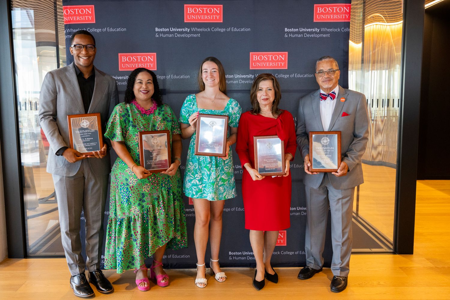 The five alumni award winners standing with their awards infront of a black step and repeat reading Boston University Wheelock College
