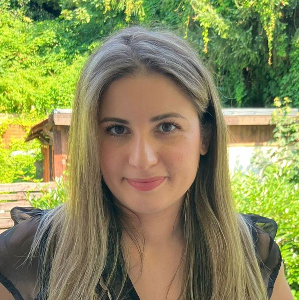 Shows Alba Franco, who is standing in an outdoor setting and has long blond hair and is wearing a black short sleeve shirt
