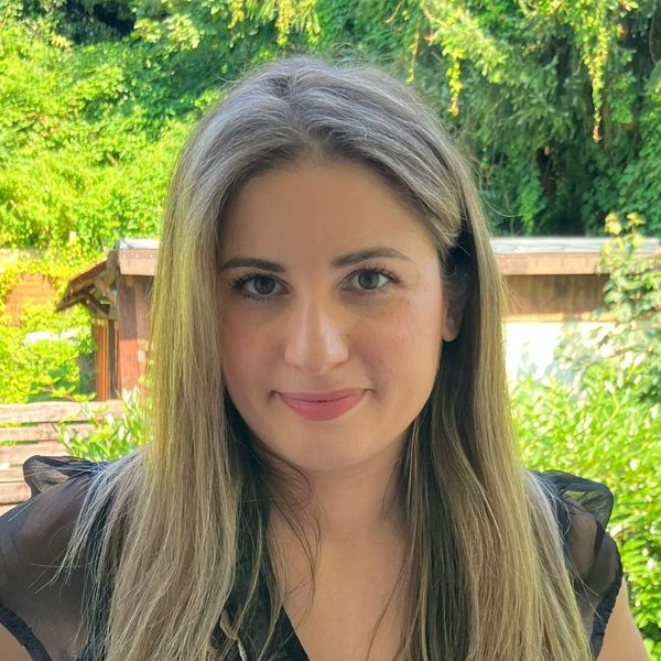 Shows Alba Franco, who is standing in an outdoor setting and has long blond hair and is wearing a black short sleeve shirt