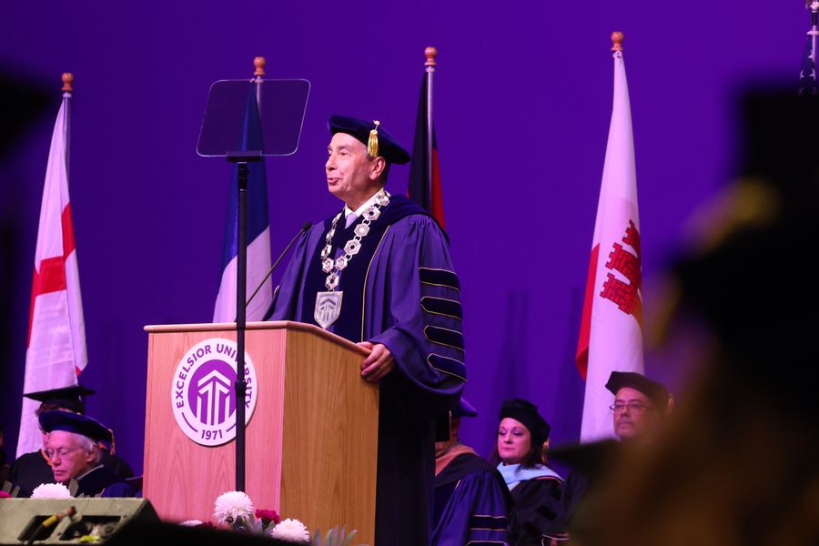 Shows a man standing at a lectern in academic regalia delivering a speech