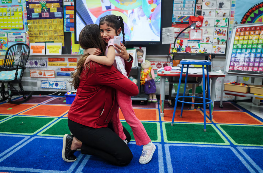 A kneeling teacher hugs a young student in a classroom