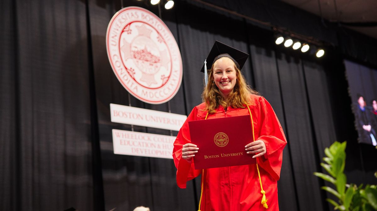 A woman dressed in a red graduation robe holds up her diploma
