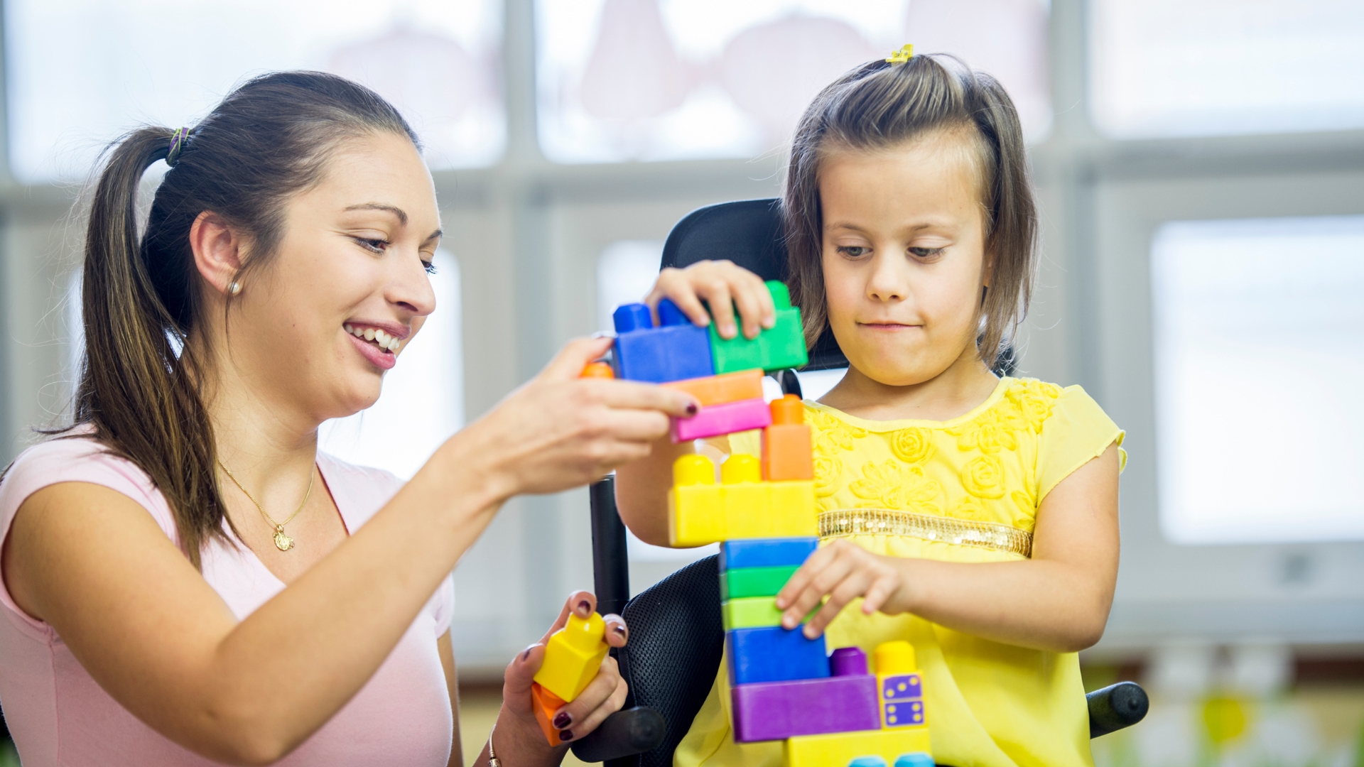 A smiling woman sits beside a young girl in a yellow dress as they stack large, colorful plastic blocks together at a table indoors.