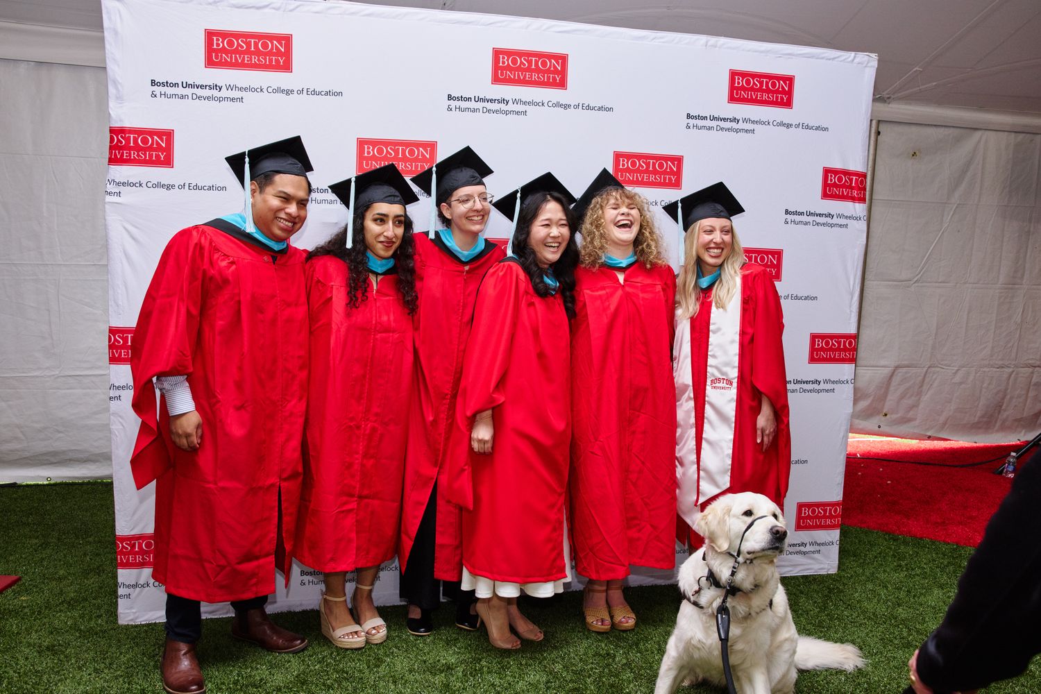 Seven graduates in red gowns and caps joyfully pose together at a Boston University event. A service dog sits calmly in front of them.