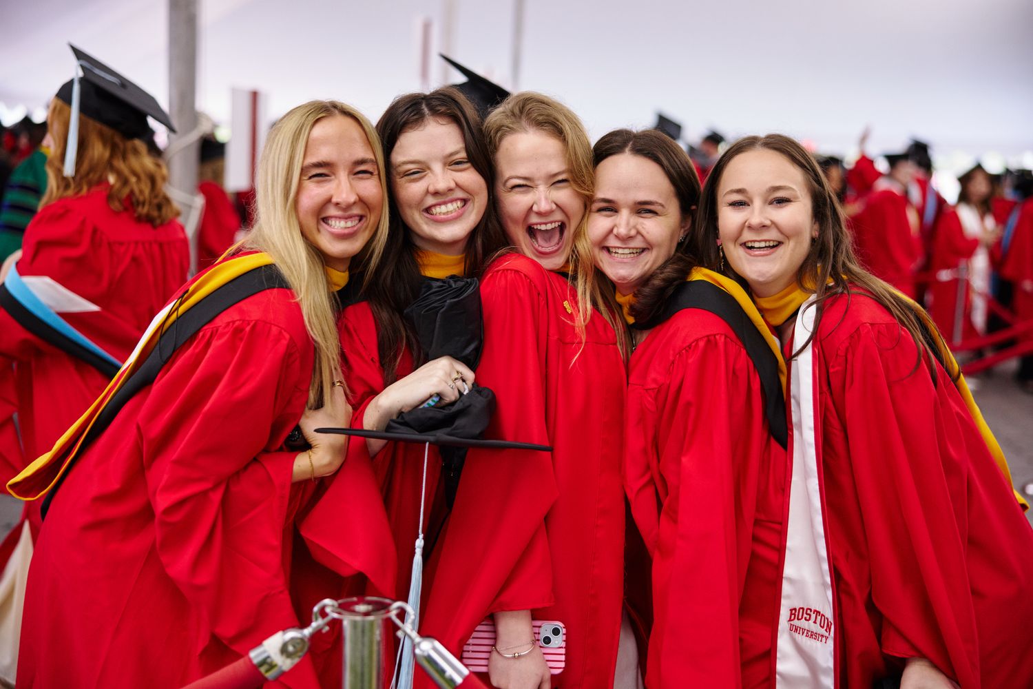 Five smiling graduates in red academic gowns pose together at a graduation ceremony under a large tent, surrounded by other students in caps and gowns.