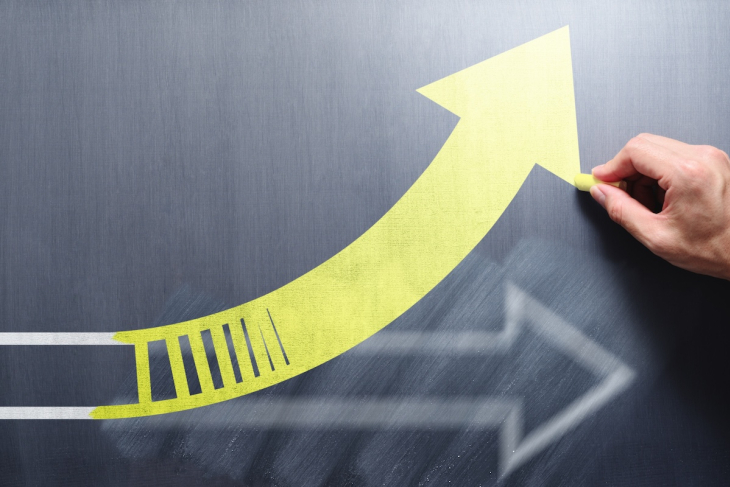 A hand draws a bright yellow arrow curving upward on a dark chalkboard, symbolizing growth and progress. A blurred white arrow points forward.