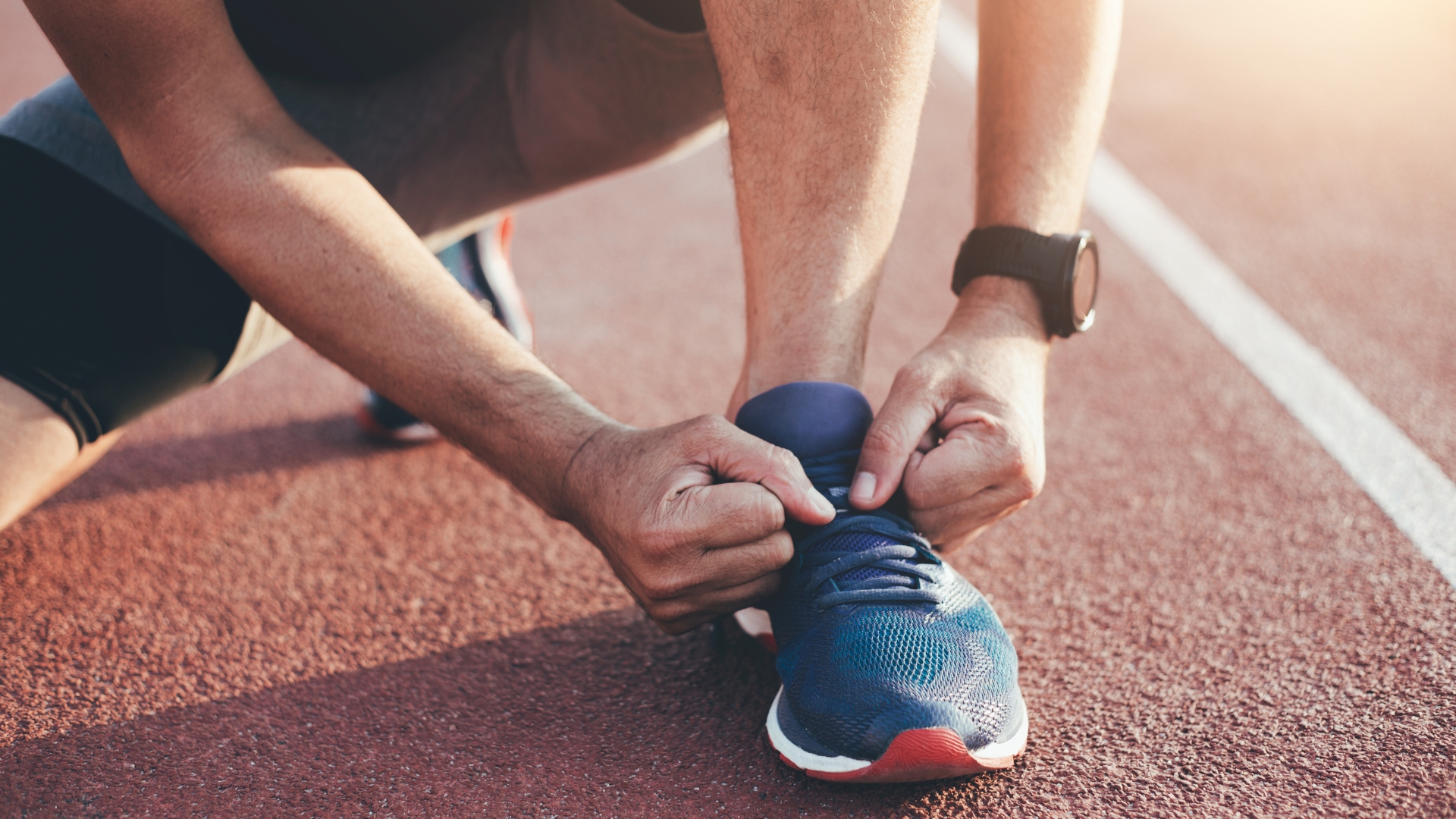 Person ties the shoelaces of a blue athletic shoe while kneeling on a red running track, wearing a black smartwatch.
