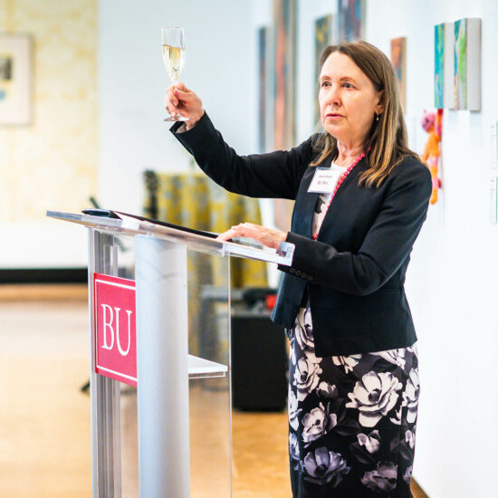 Photo: Dr. Beverly Brown, a white woman wearing a white blouse, black blazer, and black and white floral skirt, holds up a glass of champagne as she stands at a glass podium and addresses a crowd during a reception.