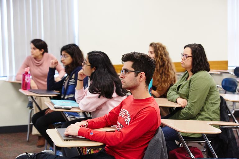 Students and instructor look sit at desks looking toward the front of the classroom.