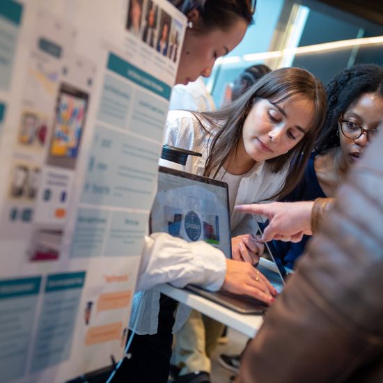 The image shows three students behind a table with a computer, next to an unidentified person in a brown jacket. They are all observing something on the screen, suggesting a moment of learning.