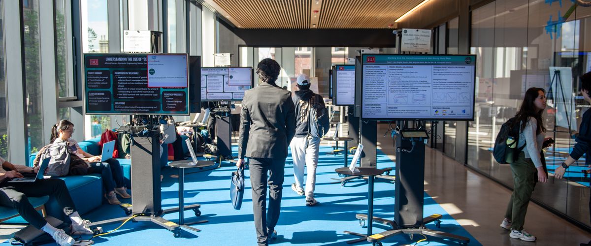 In a spacious room, screens are set up for an expo. Some students can be seen hanging around.