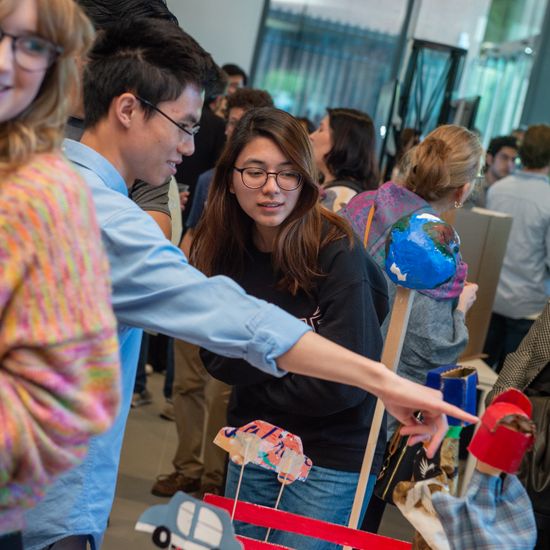 An image from a Boston University event shows a student showcasing a puppet project, flanked by two other interested students. Their various poses indicate a lively and engaging learning environment.